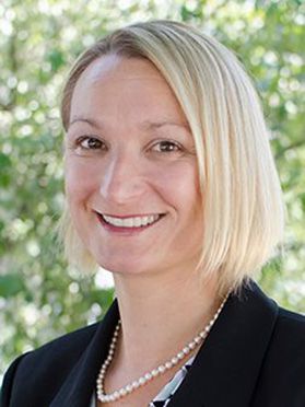 Headshot of WVU professor Kristen Matak. She is pictured outside with green trees in the background. She is wearing a black jacket over a printed blouse with a string of pearls. Her blond hair is cut in a bob. 