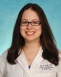 A person with long dark hair smiles while wearing black frame glasses and a white lab coat in front of a blue background. The name sewn on the white lab coat is Kacie Kidd, M.D.