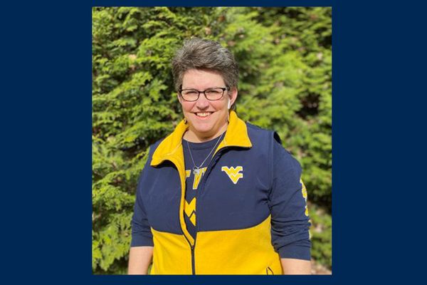 Photo of WVU doctoral student Christy Knight show here wearing a navy blue and gold WVU-branded zip up jacket over a WVU-branded T-shir. She has short salt and pepper hair and wears glasses. She is standing in front of green trees. 