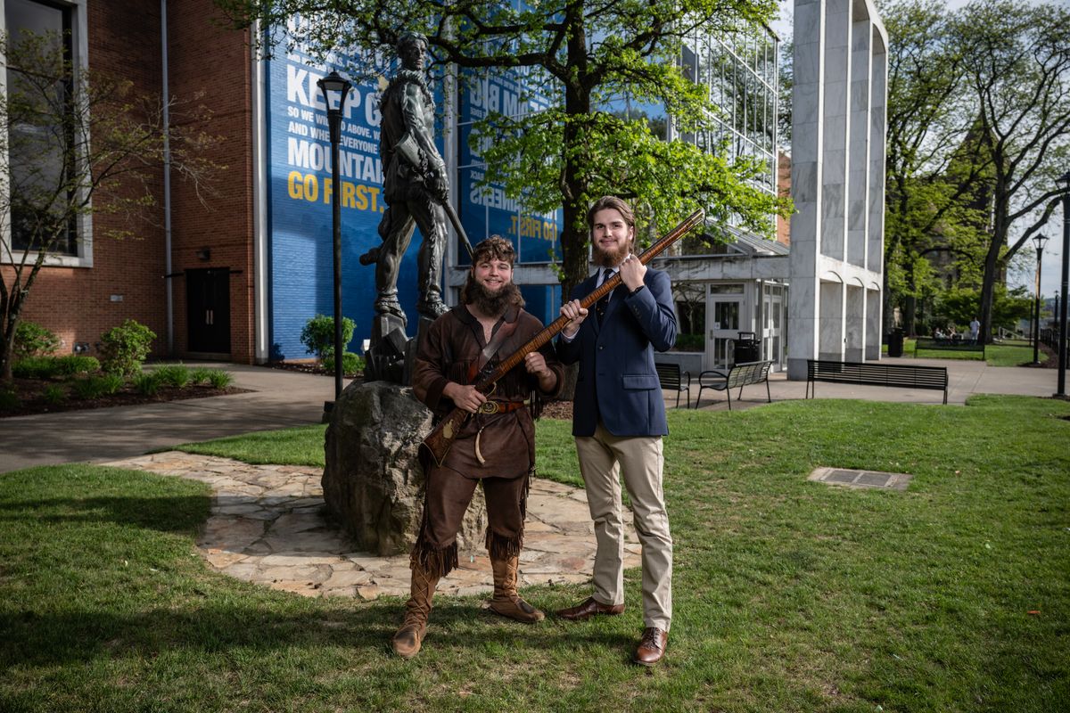 Mikel Hager, the 69th Mountaineer mascot, wears the buckskins while handing the rifle to Braden Adkins, the 70th Mountaineer mascot, outside the Mountainlair.