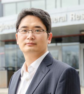 Portrait of Yuxin Wang, he has short dark brown hair and is wearing glasses and a suit.