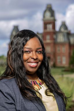Meshea Poore, Esq. is pictured here with long, dark hair, and wearing a navy blue coat, a cream colored blouse, and a colorful necklace. She is standing outside with an image of Woodburn Hall in the background. 