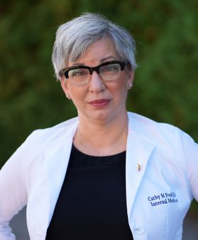 Headshot of WVU honoree Cathy Funk. She is standing outside with greenery behind her. She is wearing her white lab coat over a black T-shirt. She has very short gray hair and wears black rimmed glasses. 