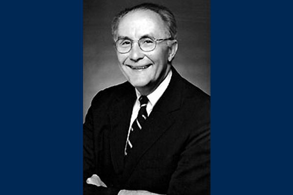 Black and white photo of Pete White, a WVU basketball standout and longtime supporter. He is pictured wearing a dark colored suit with a white shirt and striped tie. He has glasses and is smiling in the photo. 