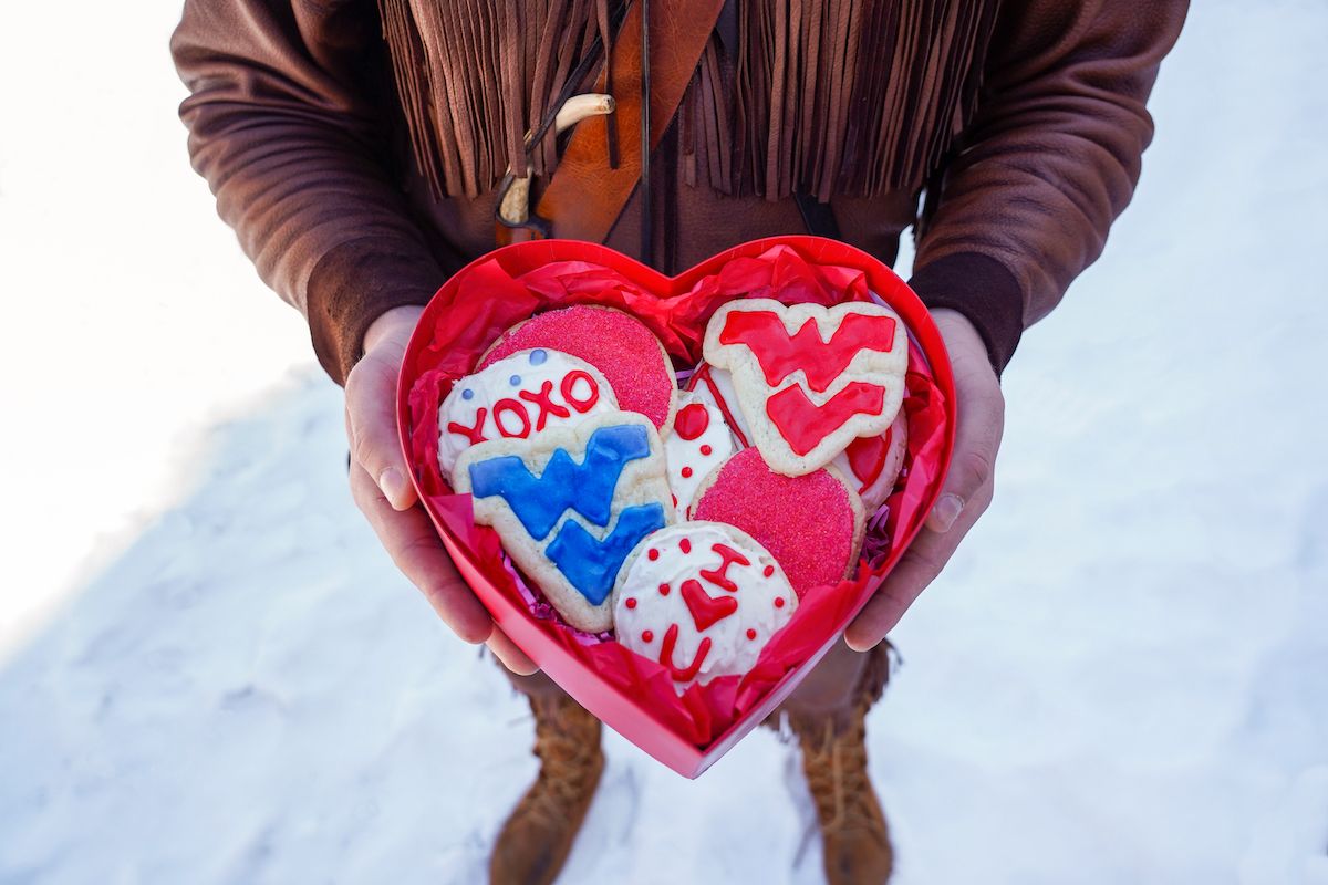 The hands of the Mountaineer mascot can be seen holding a heart-shaped tin that is open and contains more than half a dozen Valentine's Day cookies. Several have red icing. The Flying WVU is on two separate cookies - one in red and one in blue.