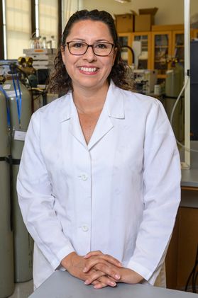 Headshot of WVU professor Tatiana Trejos. She is standing in her lab dressed in a white lab coat. Her hands are clasped together on a table in front of her. She has brown curly hair and wears glasses. 