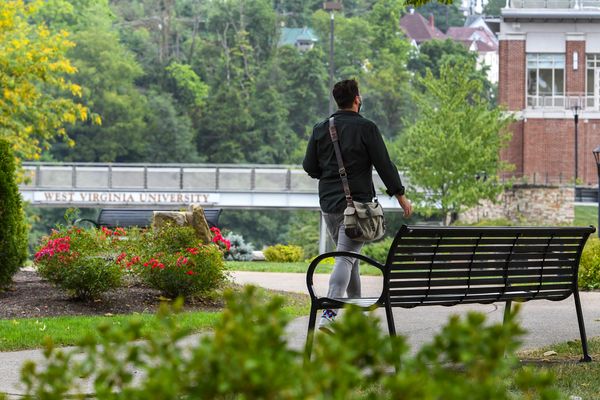 a person walks past a bench, footbridge in background, green trees, large brick building in distance
