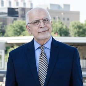 Headshot of WVU professor Samuel Ameri. Is is pictured standing outside with the engineering building complex in the background. He is wearing a navy blue sports coat with a light blue dress shirt and a striped tie. He has white hair.