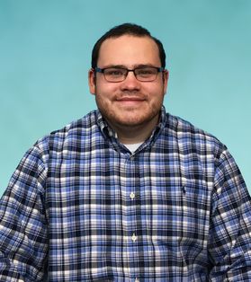 Headshot of WVU employee Cortland Nesley. He is pictured in front of a bright blue background wearing a blue and black plaid shirt. He has short dark hair and wears glasses. 