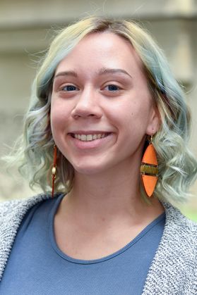 Portrait of young woman with light hair, blue-ish streaks, wearing cadet blue top and marled blue sweater with large orange earrings