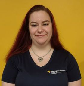 Headshot of WVU employee Sydni Vega. She is pictured standing against a gold wall and is wearing a WVU branded V-neck T-shirt. 