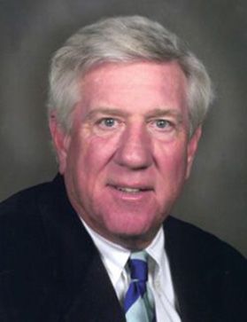 Headshot of Douglas Van Scoy. He is pictured against a gray background and is wearing a dark colored suit with a white dress shirt and a blue striped tie. He has short gray hair. 