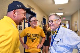 Gee stands wearing a blue shirt with a dad in a blue hat and yellow shirt and girl with dark glasses wearing yellow WVU tshirt
