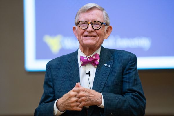 Photograph of WVU President Gordon Gee. He is wearing a navy blue suit with a white dress shirt and a pink and white polka dot bowtie. There is a presentation screen in the photo behind him. 