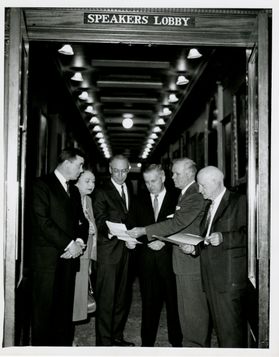 Group of people gather around a document in black and white photo.
