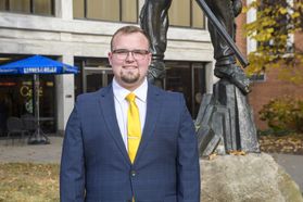 Headshot of WVU student Trevor Swiger pictured standing outside of the Mountainlair wearing a blue suit and a gold tie. 