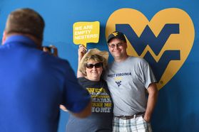 Two parents pose for a man in a blue shirt taking a picture of them in front of a blue and gold WV backdrop.