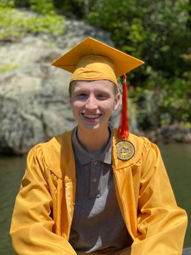 A person with short hair wears a gold cap and gown with an orange tassel over a gray polo shirt and is smiling while sitting outside in front of a rock and a body of water.