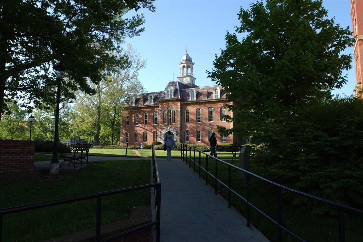 Photo of a red brick building at the end of a walkway, surrounded by shadowed trees, green grass and blue sky