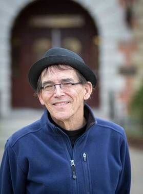 Headshot of WVU professor Joel Beeson. He is pictured outside with brick buildings in the background. he is wearing a navy blue jacket and is wearing a dark gray hat. 