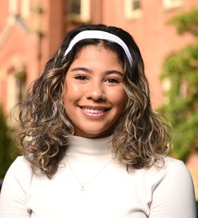 Headshot of Ariana Burks, she is pictured smiling and has curly light brown hair. She wears a white turtleneck sweater, matching the white headband in her hair. 