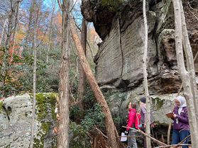 WVU Professor Vaike Haas and her students survey a new recreational spot near Morgantown, West Virginia. They are pictured in a thick forested area with giant rock cliffs in the foreground and behind them. 