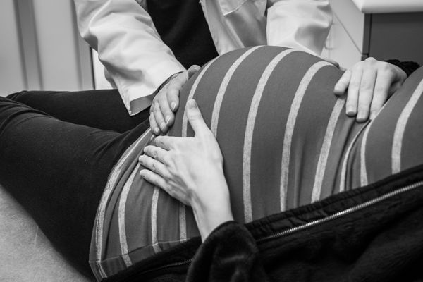 A doctor in a white coat puts two hands of the belly of a pregnant person who is dressed in dark pants and a striped top. The patient is only visible from below the neck.