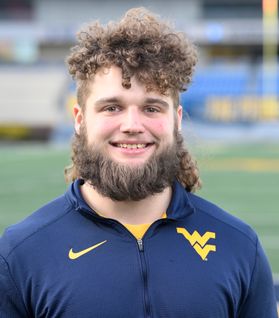 This is a portrait of Mikel Hager who has curly brown hair that sits on the top of the head and a beard. Mikel is standing on Mountaineer Field wearing a navy blue shirt with a gold Flying WV on the viewer's right side.