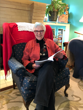 Photo of Judith Stizel. She is seated in a navy blue patterned chair and is holding a manuscript. She is wearing black pants with a black shirt and red jacket. She has short gray hair. 