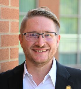 Headshot of Sam Taylor. He is pictured standing outside in front of a red brick building with windows. He is wearing a dark colored suit jacket over a white dress shirt with a gold lapel pin. He has short blonde hair, a light beard and wears glasses. 