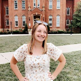 Photograph of WVU student Jordan Murray. She is standing in Woodburn Circle with her hands on her hips. She is wearing a floral dress and has sunglasses perched on top of her head. She also has long, dirty blonde hair. 