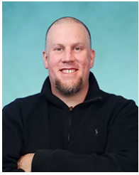 Sam Wilkinson, program manager for Regional Transition Navigator, poses for a headshot wearing a dark quarter zip shirt. He is bald with a goatee, and his arms are crossed. 