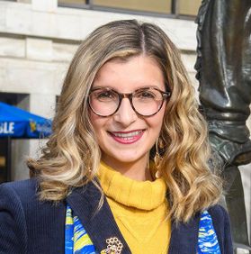 This is a portrait of Giana Loretta who is shown outside the Mountainlair in front of the Mountaineer statue and is wearing a gold sweater, blue jacket and gold and blue scarf.