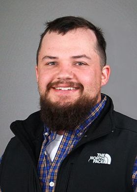A man with short brown hair and a beard. He is wearing a black The North Face jacket over a blue flannel.