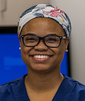 A woman wearing glasses and a flowery bandana on her head smiles in blue scrubs.