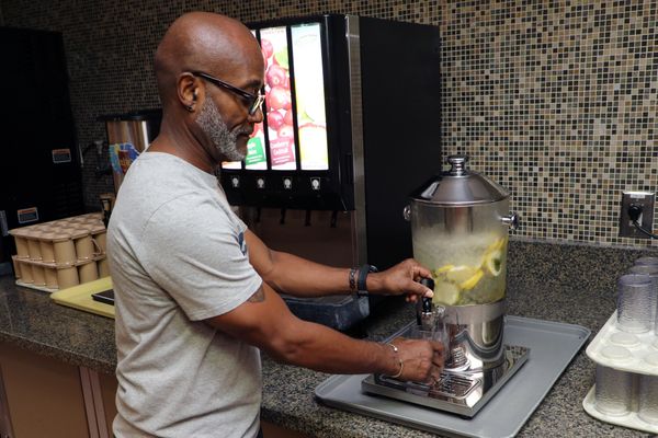 A gentleman with a grey beard and glasses in a grey t-shirt fills his cup with lemon-infused water.