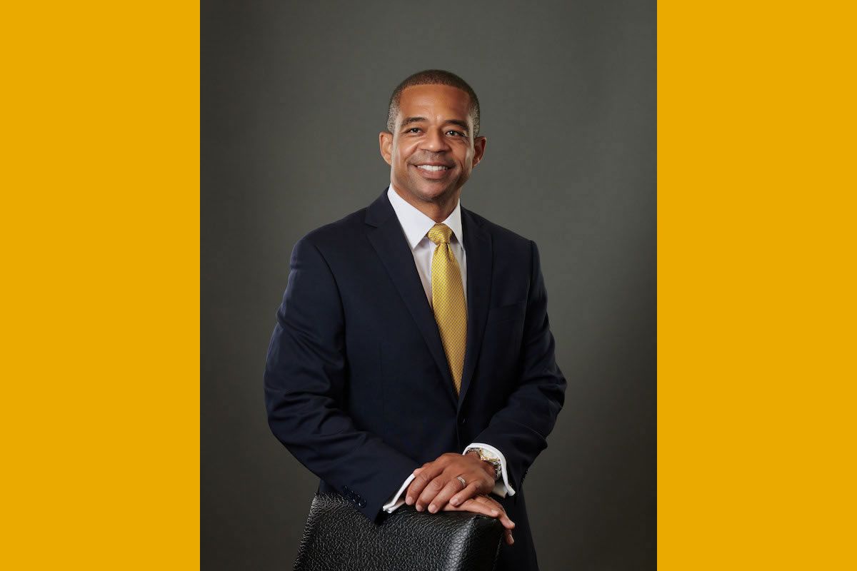 Headshot of T. Ramon Stuart, the new president of WV Institute of Tech. Gold is standing with his hands crossed on a chair. He is wearing a dark colored suit and a gold tie. He is a Black man with short hair, and is smiling slightly in the photo. 