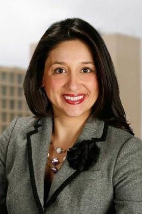 Headshot of WVU administrator Stephanie Taylor. She is pictured inside wearing a gray tweed jacket with a black floral label pin and silver statement necklace. She has long black hair and wears red lipstick. 