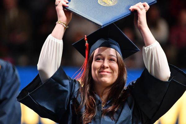 A WVU graduate holds a blue diploma folder above their head while wearing a navy blue graduation gown.
