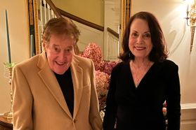 William and Patricia Bright stand side by side in a beige hallway. William is wearing a light brown jacket over a dark shirt. Patricia has on a black v-neck top and shoulder-length dark hair. 