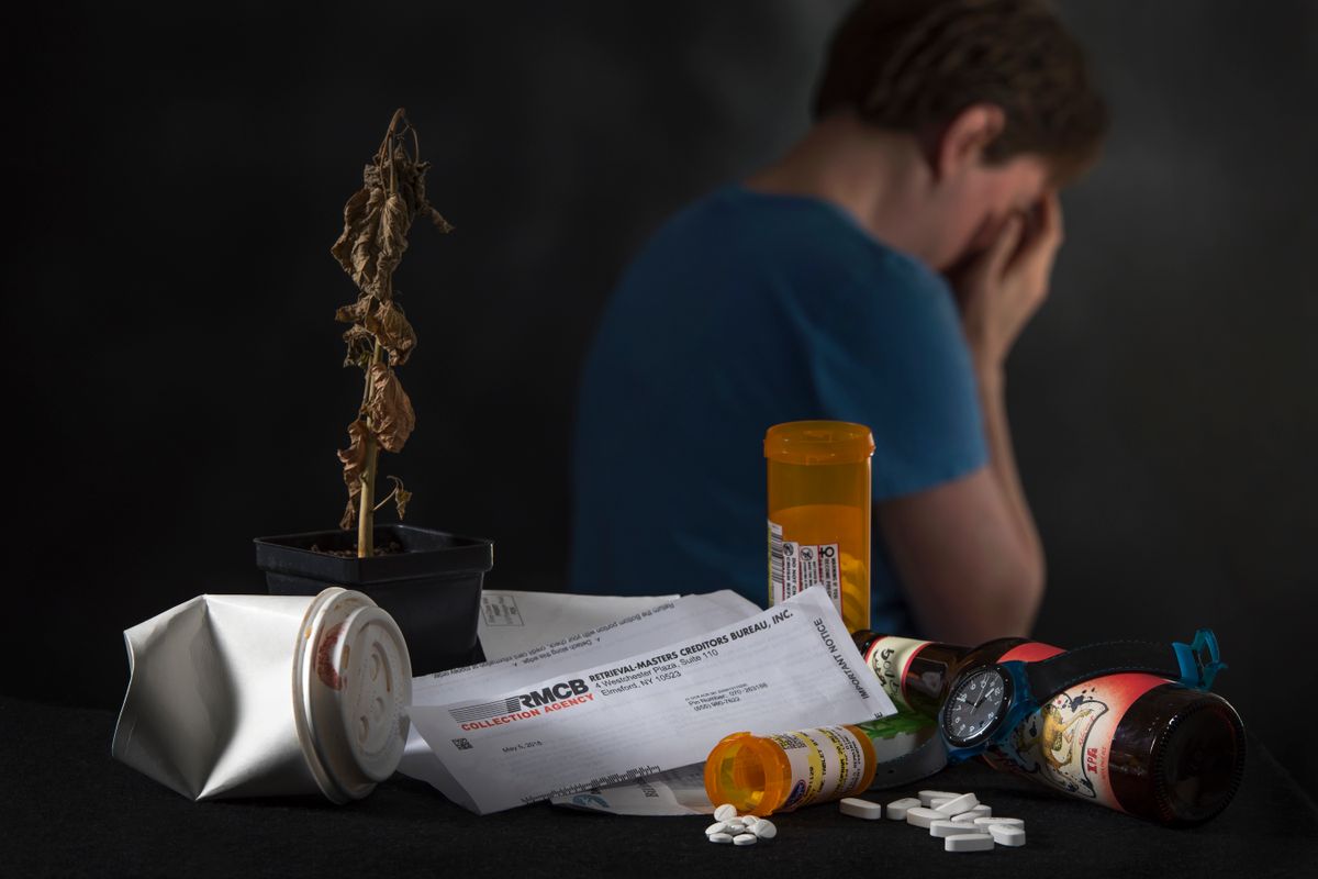 A man sits with his face in hands, his back to a pile of mail and opened pill bottles