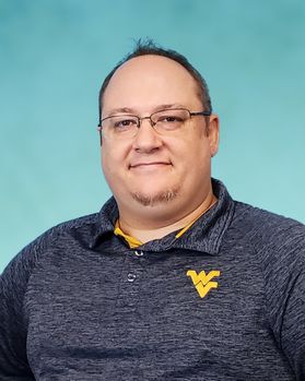 Headshot of Doug Cumpsten. He is pictured in front of a light blue wall and is wearing a heathered navy blue golf shirt with a Flying WV on the pocket. He has very short brown hair and wears glasses. 