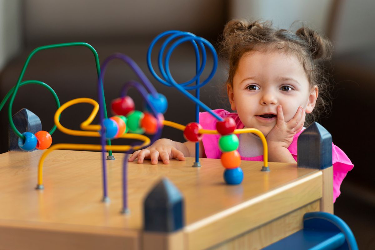 An image showing a young girl playing with a toy in a doctor's office waiting room. The toy is brightly colored with beads on wires that can be moved about. The girl has brown hair and is wearing a pink shirt. 