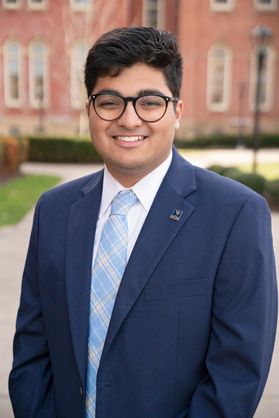 A WVU student wearing a dark blue suit jacket over a white button up shirt and a light blue tire. He has dark hair with round glasses.