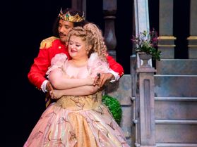 A female actress in a pink dress performs in a scene with a male actor wearing a red coat and a gold crown. 