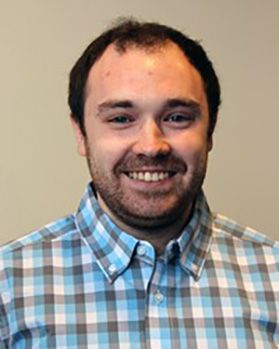 Headshot of WVU graduate student James Scripter. He is standing in front of a beige wall wearing a blue, gray and white plaid shit. He has short dark hair and a light beard. 