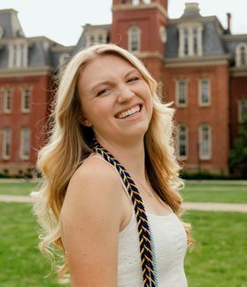 Headshot of WVU student Erin Langan. She is pictured outside in front of Woodburn Hall. She is wearing a white top with braided chords around her neck. She has long blonde hair. 