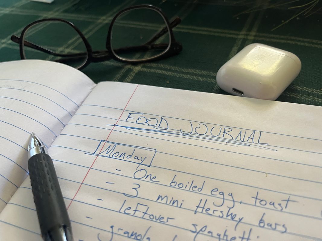 A photograph of a food journal which includes items written in blue ink on a notebook. There are also glasses, a pen and a airpods case sitting around the notebook. 