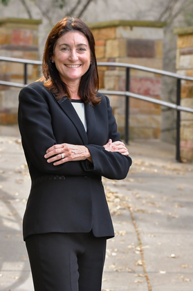 Headshot of associate professor Carla Brigandi. She is standing outside with her arms crossed. She is wearing a black pant suit and white blouse. She has medium length brown hair/