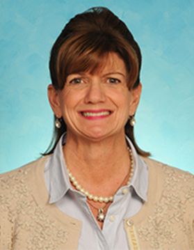 A smiling woman with beige jacket, gray shirt, pearls on bright blue background